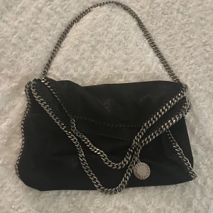 Svart axelväska med kedjedetaljer - Snygg svart axelväska med silverfärgade kedjor som axelrem och kantdetaljer. Perfekt för att ge en edgy touch till din outfit. Väskan har en rund metallbricka som dekoration.