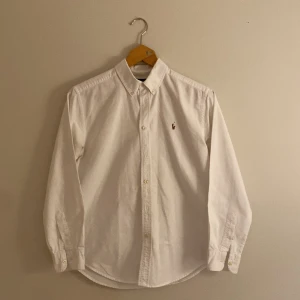Vit skjorta från Ralph Lauren - Klassisk vit skjorta från Ralph Lauren med broderad logotyp på bröstet. Skjortan har långa ärmar och knappar framtill. Perfekt för en stilren look.