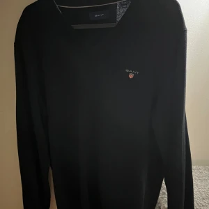 Svart tröja från GANT - Säljer en stilren svart tröja från GANT med v-ringning och broderad logotyp på bröstet. Perfekt för en klassisk look. Endast testad så skicket är som nytt , storlek M men passar även S 