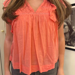 Korallfärgad blus med volangdetaljer - Säljer en söt korallfärgad blus med volangdetaljer och korta ärmar. Blusen har en lös passform och v-ringad hals. Perfekt för vår och sommar!