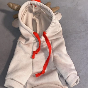 Beige huvtröja med horn och röda detaljer till hundar  - Söt beige huvtröja med horn och röda snören. Perfekt för din hund. Tröjan har en mysig huva och är gjord i ett mjukt material. Passar även till julen. Den är helt ny använd 1 gång.Säljer på grund av den har blivit för liten för min hund. står inte vad det är för storlek men tror att det är XS/S😊