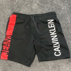 Svarta shorts från Calvin Klein - Snygga svarta shorts från Calvin Klein med röd och vit textdetalj på sidan. Shortsen är köpta från Kids Brand Store och är använda ett par gånger men är i bra skick. Dom är i storlek S. 