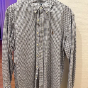 Rutig skjorta från Ralph Lauren - Klassisk rutig skjorta från Ralph Lauren i blått och vitt. Skjortan har långa ärmar och knappar framtill samt en broderad logotyp på bröstet. Perfekt för en stilren look.