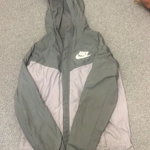 Grå och vit vindjacka från Nike - Säljer en stilren grå och vit vindjacka från Nike. Jackan har en huva och dragkedja framtill, perfekt för blåsiga dagar. Den är lätt och smidig att bära med sig. Nike-loggan syns tydligt på bröstet.