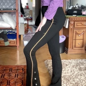 Svarta ställbara byxor med knappar - Snygga svarta byxor med en orange linje längs sidan och knappar vid bensluten.  Längd: 104cm  Bredd: 35cm