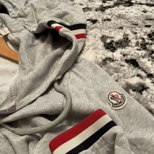Grå huvtröja från Moncler - Snygg grå huvtröja från Moncler med dragkedja och randiga detaljer i rött, vitt och svart på ärmar och huva. Perfekt för en avslappnad stil med en touch av lyx. Har inget kvitto på plagget, men kan säkert lösa något.