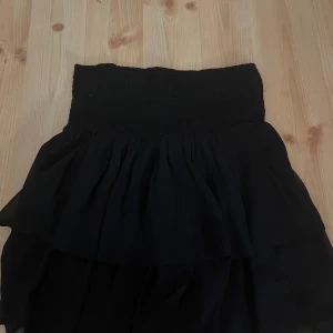 Svart kjol med volang - Snygg svart kjol med volangdetaljer och elastisk midja. Perfekt för en stilren look. Använd fåtal gånger.