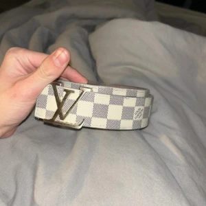 Bälte - Snyggt bälte med ett klassiskt rutigt mönster i beige och grått. Bältet har ett stilrent monogramspänne i silverfärg. Perfekt för att ge en touch av elegans till din outfit.