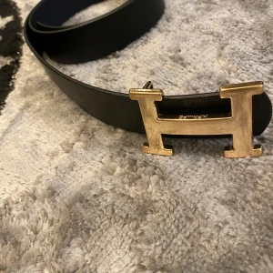 Svart bälte med guldfärgat H-spänne - Snyggt svart bälte i skinn med ett iögonfallande guldfärgat H-spänne. Perfekt för att ge en lyxig touch till din outfit. Passar till både jeans och kostymbyxor.