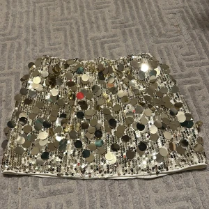 Glittrig paljettkjol - Snygg och iögonfallande kortkjol täckt med glittriga paljetter i silver. Perfekt för att sticka ut på festen. Kjolen har en elastisk midja för bekväm passform.