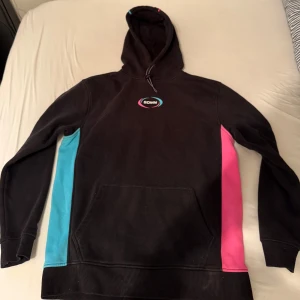 Svart hoodie från Sidemen - Säljer en svart hoodie från Sidemen (YouTube) med färgglada detaljer i rosa och blått på sidorna. Den har en stor ficka framtill och justerbar huva med snören. Perfekt för en avslappnad stil.