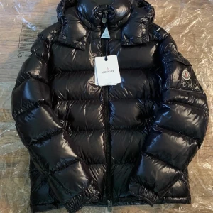 Svart dunjacka från Moncler - Snygg och glansig svart dunjacka från Moncler med huva och dragkedja. Perfekt för kyliga dagar med sin pufferdesign och långa ärmar. Moncler-logga på ärmen ger en exklusiv touch. Använd i gott skick endast seriösa köpare pris kan diskuteras vid snabb affär pm vid fler bilder