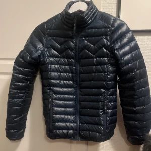 Mörkblå dunjacka från Mountain Works - Snygg mörkblå dunjacka från Mountain Works med quiltad design och rosa insida. Jackan har en hög krage och är perfekt för kyligare väder. Den är i väldigt bra skick, endast använd några få gånger. Nypris 1995 kr.