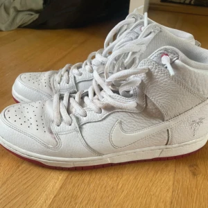 Vita Nike Dunk sneakers - Snygga vita Nike Dunk sneakers med hög design och perforerade detaljer på tån. Skorna har en röd sula och klassisk snörning. Insidan har ett unikt tryck med palmmotiv. Perfekta för en stilren look.