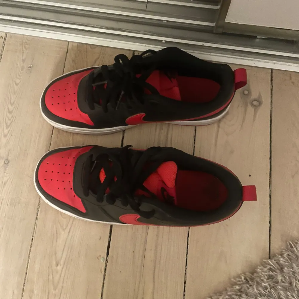 Snygga Nike sneakers i svart och rött med klassisk design. Skorna har en röd Nike-logga på sidan och svart snörning. . Kengät.