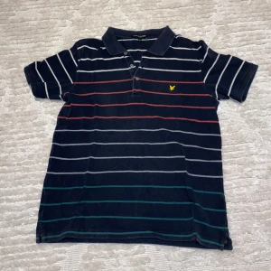 Randig pikétröja från Lyle & Scott - Snygg randig pikétröja från Lyle & Scott i marinblått med vita, röda och gröna ränder. Tröjan har korta ärmar och en klassisk krage med knappar. Perfekt för en avslappnad stil.