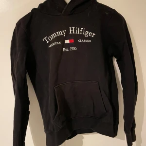 Svart hoodie från Tommy Hilfiger - Säljer en klassisk svart hoodie från Tommy Hilfiger med tryck på framsidan. Hoodien har en känguruficka och en bekväm passform. Perfekt för en avslappnad stil.