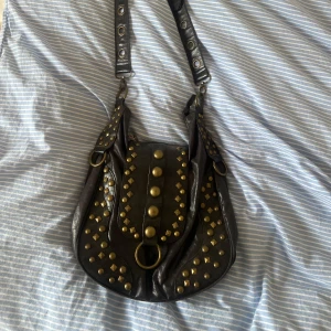Svart axelväska med nitar - Snygg svart axelväska i skinn med coola nitar i guldton. Väskan har en justerbar axelrem och flera dekorativa detaljer.