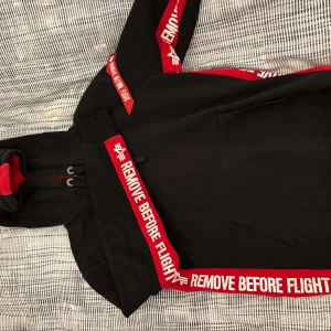Svart hoodie med röd text från Alpha industries  - Snygg svart hoodie med röd text 'REMOVE BEFORE FLIGHT' längs ärmar och huva. Hoodien har en praktisk ficka framtill och justerbar huva med snören. Perfekt för en avslappnad stil.
