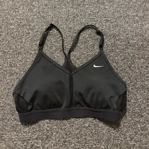 Svart sport-bh från Nike - Säljer en svart sport-bh från Nike med justerbara axelband och en liten vit logga på framsidan. Perfekt för träning och ger bra stöd. Tillverkad i ett mjukt och stretchigt material för optimal komfort.