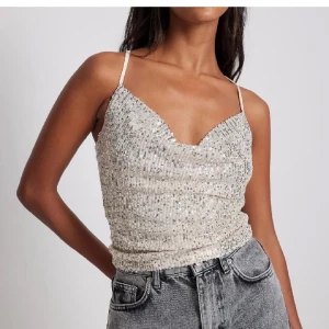 Glittrig linne i silver - Snygg glittrig linne i silver med smala axelband och draperad framsida. Perfekt för en festlig kväll. Axelbanden korsas i ryggen för en extra detalj.