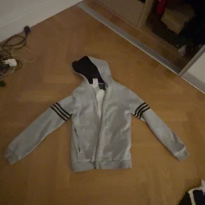 Grå huvtröja från Adidas - Säljer en grå huvtröja från Adidas med svarta ränder på ärmarna och en stor logga på ryggen. Tröjan har dragkedja. Den är ganska använd och har därför små hål här o där och en liten fläck.