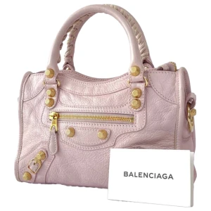 Ljusrosa mini city bag från Balenciaga - Säljer en elegant ljusrosa handväska från Balenciaga i skinn. Väskan har guldfärgade detaljer, flätade handtag och en rymlig insida med svart foder. Köpt från Vestiere Collective 🩷Endast använd en gång sedan  köpt från V.C 💛