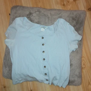 Ljusblå  kortärmad topp från Monki - Säljer en ljusblå  kortärmad topp från Monki med knappar framtill. Toppen har en rund halsringning och är tillverkad i ett mjukt material med diskret mönster.