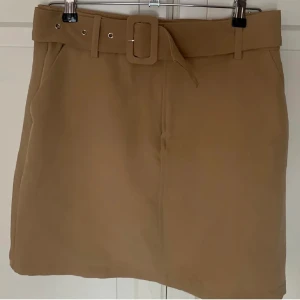 Beige kjol med bälte - Snygg beige kjol med ett matchande bälte i midjan. Kjolen har en stilren design och är perfekt för en chic look. Den har en normal passform och är tillverkad i ett mjukt material.