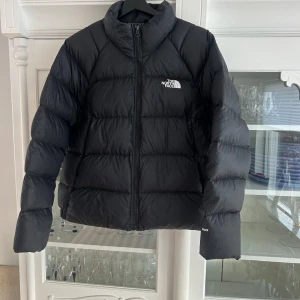 Svart dunjacka från The North Face - Säljer en svart dunjacka från The North Face med dragkedja och hög krage. Jackan har en pufferdesign och är perfekt för kyligare väder. Logotypen är tryckt på bröstet och ryggen. Från djur & rökfritt hem. 