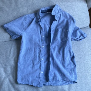 Blå kortärmad skjorta från H&M - Säljer en blå, jättesnygg kortärmad skjorta från H&M. Perfekt för varma dagar och avslappnade tillfällen. Klassisk, snygg Stockholmsstil och skjortan är as fin till killar och storlek 146