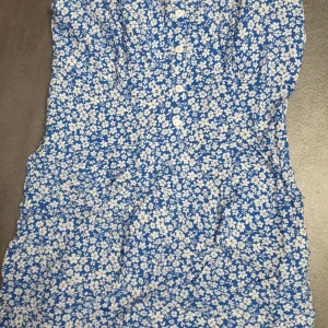 Blå blommig klänning - Charmig blå klänning med vita blommor och knappar framtill. Perfekt för en somrig look.