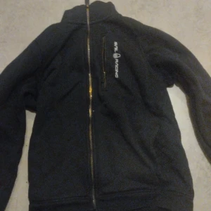 Svart fleecejacka från Sail Racing - Säljer en svart fleecejacka från Sail Racing med dragkedja framtill. Jackan har en hög krage och en bröstficka med dragkedja. Perfekt för kyliga dagar.