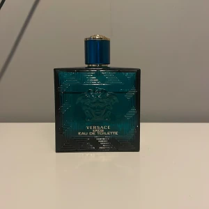 Versace Eros Eau de Toilette - Säljer en flaska Versace Eros Eau de Toilette. Flaskan är fyrkantig och har en djup turkos färg med ett elegant mönster. Den har en blå kork med gulddetaljer och den ikoniska Medusa-loggan på framsidan. Perfekt för den som vill ha en doft med karaktär och stil.