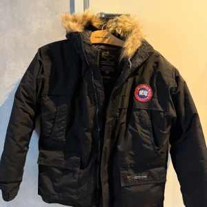 Svart vinterjacka från Canada Goose - Säljer en svart vinterjacka från Canada Goose med pälsfodrad huva och broderad logotyp på bröstet. Jackan har dragkedja och knappar framtill samt flera praktiska fickor. Perfekt för kalla vinterdagar.