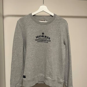 Grå Morris sweatshirt - Säljer en stilren grå sweatshirt från Morris med tryck på bröstet. Tröjan har långa ärmar och en rund halsringning. Perfekt för en avslappnad stil.