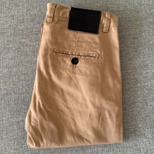 Beige chinos från W.BKTR - Snygga beige chinos från W.BKTR i storlek 30. Tillverkade i en bomullsblandning med 3% elastan för extra komfort. Klassisk design med knappar och fickor både fram och bak. Perfekta för en stilren look.