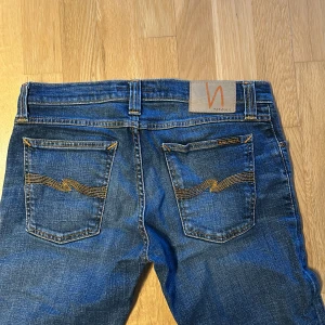Blå jeans från Nudie Jeans - Snygga blå jeans från Nudie Jeans med klassisk femficksdesign och orange sömmar. De har en knappgylf och är i en smal passform. Perfekta för en stilren look. Typ oanvända. Bra skick. 