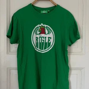 Grön t-shirt med Rögle-logga - Säljer en grön t-shirt med Rögle-logga på bröstet. T-shirten är kortärmad och har en rund halsringning. Perfekt för sportiga tillfällen eller för att visa ditt stöd för laget. Ett lagat hål 