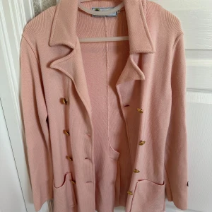 Busnel Rosa kappa med knappar  - Snygg rosa kappa med guldknappar och två fickor framtill. Perfekt för en stilren look. Den har en klassisk krage och är långärmad.