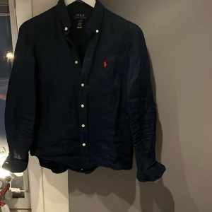 Mörkblå skjorta från Ralph Lauren - Snygg mörkblå skjorta från Ralph Lauren med klassisk krage och knappar framtill. Den har en broderad röd logotyp på bröstet och är tillverkad i ett bekvämt material. Perfekt för en stilren look.