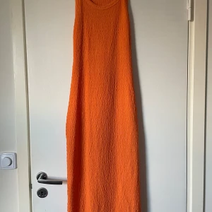 Orange ärmlös långklänning - Snygg och enkel orange långklänning med en texturerad yta. Klänningen är ärmlös och har en öppen rygg, vilket ger en luftig och somrig känsla. Perfekt för varma dagar eller kvällar.
