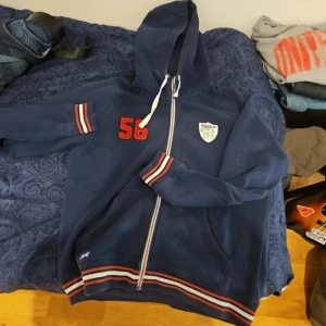 Blå hoodie med dragkedja och broderad detalj - Säljer en blå hoodie med dragkedja och huva. Den har röda och vita ränder vid ärmslut och nederkant samt en broderad detalj med siffran 56 på bröstet. Perfekt för en avslappnad stil.