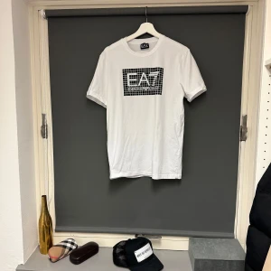 Vit t-shirt från EA7 - Säljer en snygg vit t-shirt från EA7 med ett coolt rutmönster och logga på bröstet. T-shirten är kortärmad och gjord i mjuk bomull för bästa komfort. Jättebra skick, ej sliten i tryck eller material alls! Storlek M. 