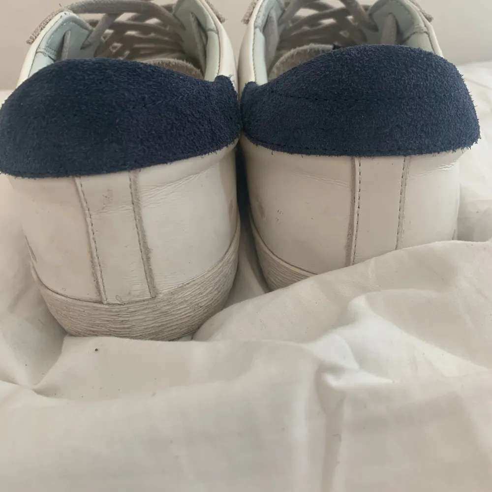 Säljer ett par vita sneakers från Golden Goose med snygga blå detaljer. De har en cool stjärna på sidan och är i välanvänt skick. Perfekta för en avslappnad stil och passar till både jeans och shorts. Skorna har snörning och är gjorda i skinn med mockadetaljer.                                                    Hör av er vid funderingar.. Skor.