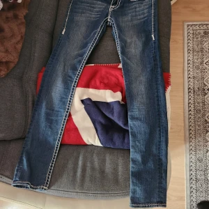 Blåa lågmidjade jeans [True Religion] - Säljer ett par snygga blå jeans från True Religion i storlek 28. De har en klassisk femficksdesign med vita sömmar och broderade bakfickor. Perfekta för en casual look eller en utekväll. Made in USA och i bra skick! Har inte använts men är väldigt lågmidjade och inte nån stretch alls. Har du större "bakredel" eller lår så köp inte.