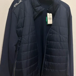 Mörkblå jacka från Ralph Lauren - Säljer en snygg mörkblå jacka från Ralph Lauren i nyskick. Jackan har en quiltad design framtill med dragkedja och RLX-logga på axeln. Perfekt för höst och vår, med långärmade ärmar och en stilren look. Passar både till vardags och lite finare tillfällen.