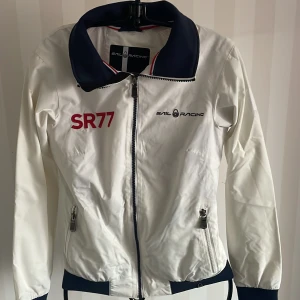 Vit jacka från Sail Racing Ny - Säljer en snygg vit jacka från Sail Racing med SR77-tryck och brittisk flagga på ryggen. Jackan har dragkedja och två fickor med dragkedjor. Perfekt för vår och höst, med en sportig stil. Den är i ny och passar perfekt för segling eller andra utomhusaktiviteter.