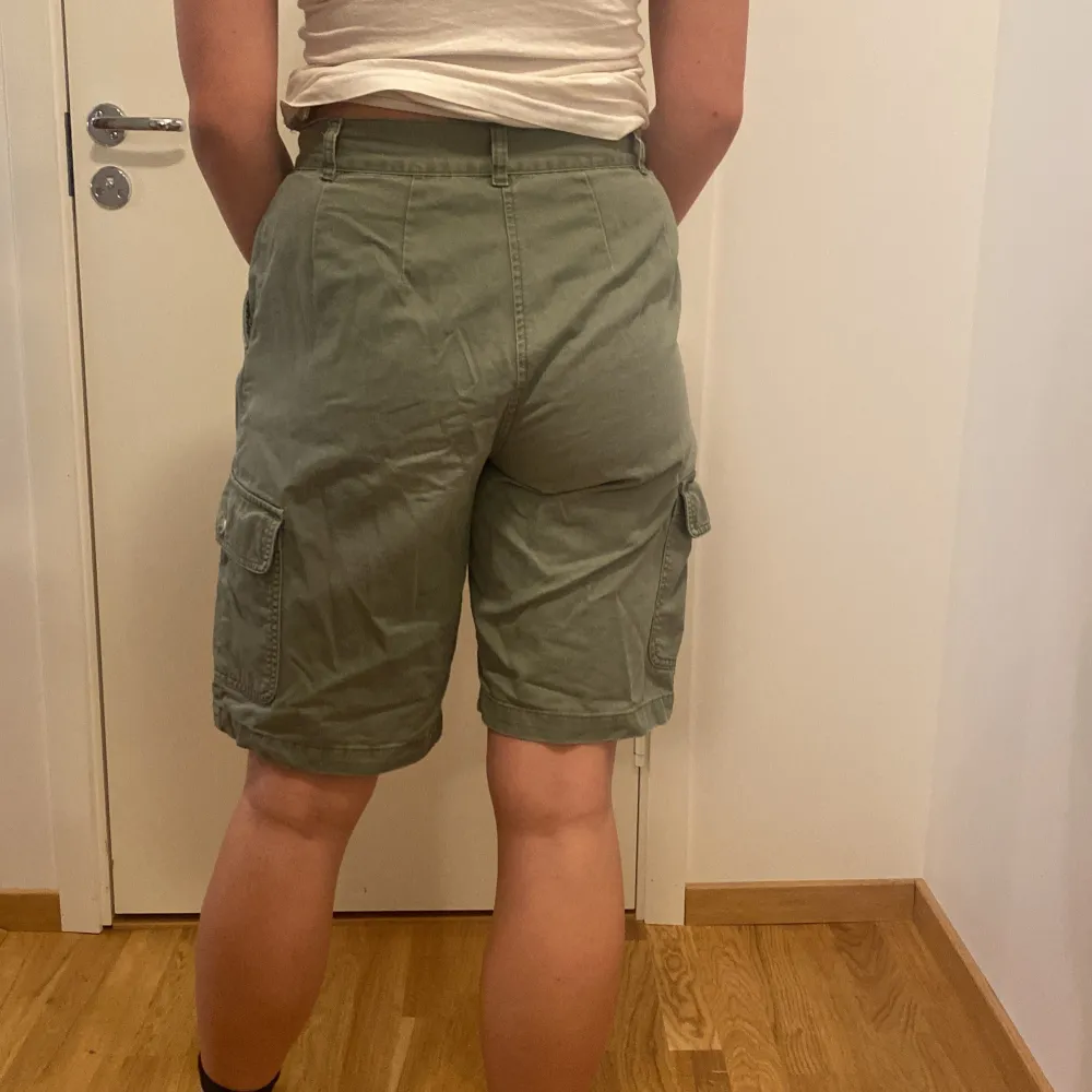 Säljer ett par gröna cargo shorts i bomull. De har flera praktiska fickor med knappar och en bekväm passform. Perfekta för sommarens äventyr eller en avslappnad dag ute. Shortsens stil är både funktionell och trendig. Uppskattad storlek M. Fattas en knapp på fickan.. Shorts.