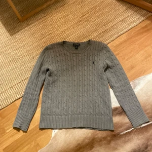Grå stickad tröja från Ralph Lauren - Säljer en stilren grå stickad tröja från Ralph Lauren med kabelstickat mönster. Tröjan har långa ärmar och en broderad logotyp på bröstet. Perfekt för kyligare dagar!